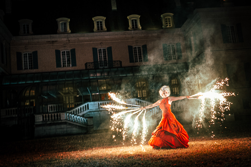 Devant un château, la comédienne vêtue d’une grande robe à volant manie deux éventails de feux d’artifice. Ces derniers forment comme des ailes lumineuses de chaque côté de l’artiste. Leur lumière illumine la robe de la comédienne ainsi que la façade du bâtiment. 