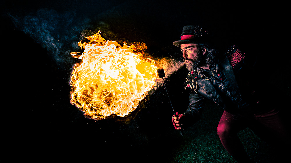 A droite, le comédien accroupi et torche en main crache une boule de feu vers le centre de l’image. La lumière chaude de la flamme éclaire le visage de l’artiste ainsi que son costume. Il porte une queue de pie noire aux épaulettes à plumes noires et blanches et orné de quelques roses du même rouge que son pantalon. Sur sa tête trône un chapeau noir entouré d’un ruban rouge. Image tirée du spectacle Kissed by Fire de la Cie Médusa (Grenoble). 