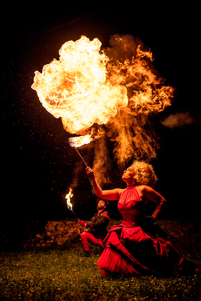 Au premier plan, la comédienne vêtue d’une longue robe rouge à volant est accroupie. Elle tient devant elle une torche et crache une boule de feu au-dessus de sa tête, en direction du centre de l’image. Au second plan, le comédien, vêtue d’une queue de pie noire, d’un pantalon rouge et d’un chapeau, regarde sa collègue exercer son art. Image tirée du spectacle Kissed by Fire de la Cie Médusa (Grenoble).